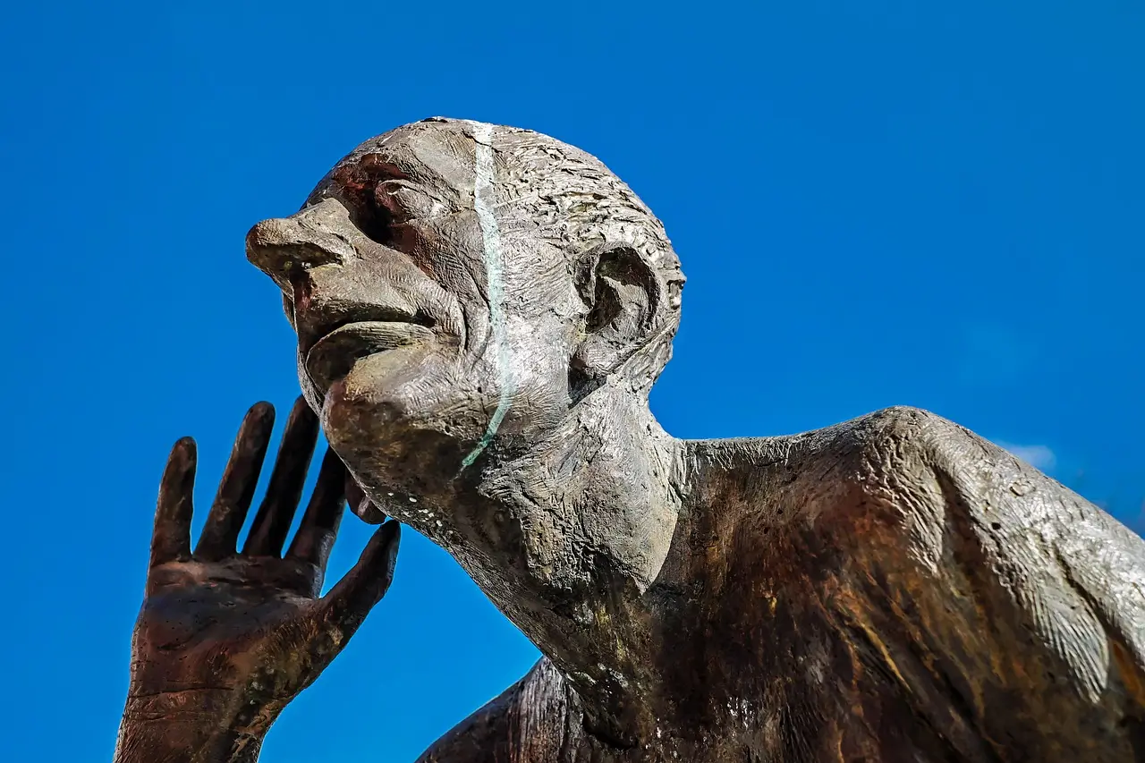 Sculpture représentant un homme en posture d'écoute, avec une main près de l'oreille, sur fond de ciel bleu.